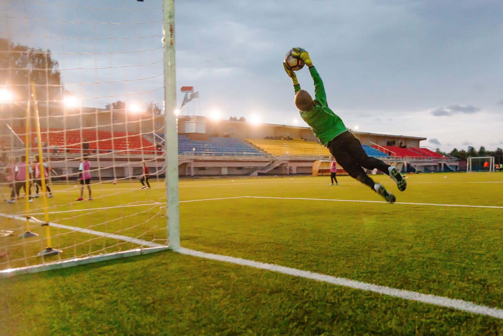A young goalkeeper at full stretch under the floodlights, catching the ball at evening training